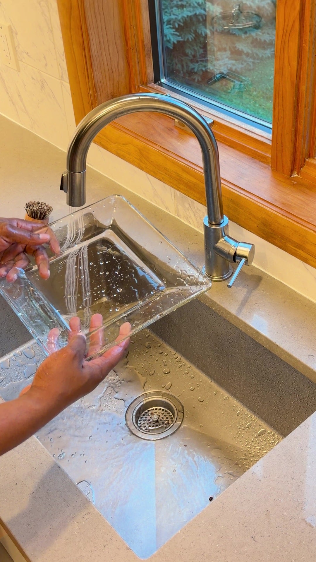 A rinsed glass dish held under the faucet at a kitchen sink by the window